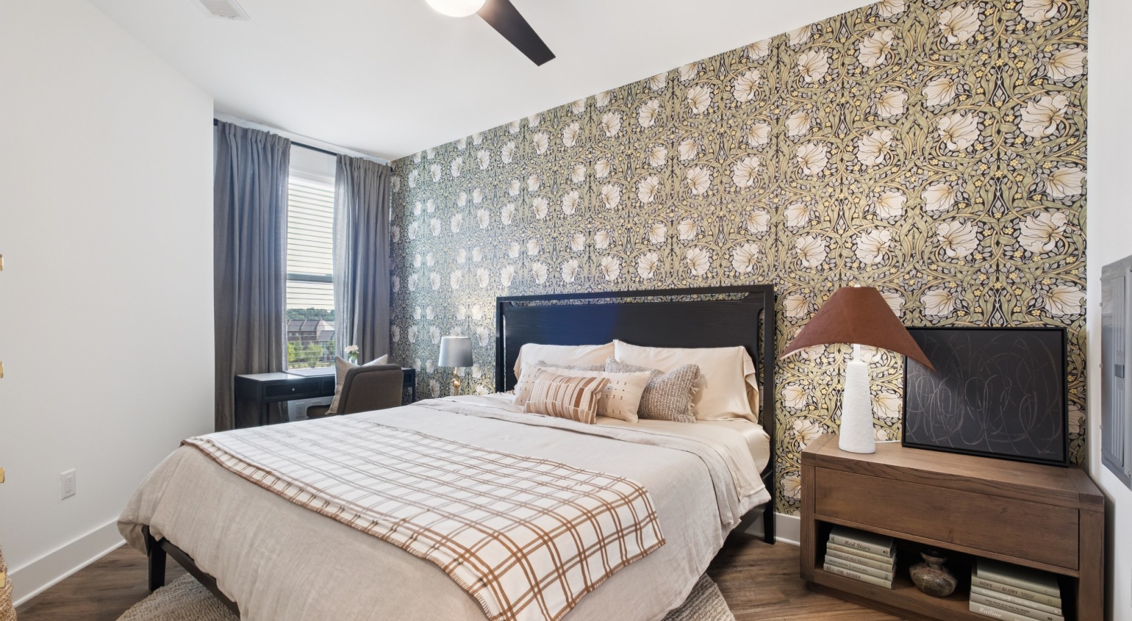 Chic bedroom with stylish floral wallpaper, elegant ceiling fan, and cozy bedding set, featuring a desk by the window with natural light in a modern apartment at The Rowley in Murfreesboro.